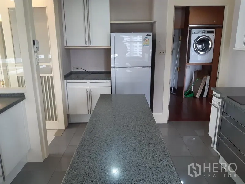 Condominium for rent in Khlong Toei, Bangkok - Kitchen island facing a refrigerator with doorway to a separate laundry room fitted with a stacked washer and dryer.