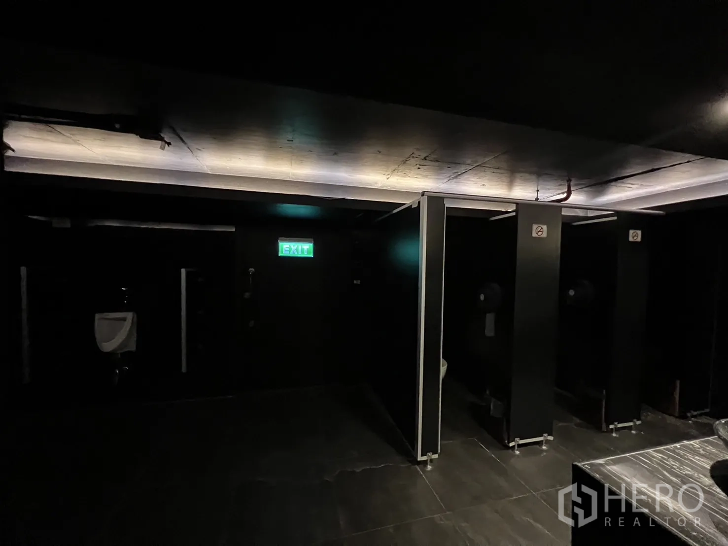 Retail Space for rent - Restroom corridor with stalls, a urinal, and an illuminated exit sign.