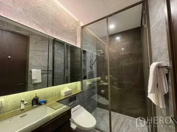 Condominium for rent in Khlong Toei, Bangkok - Modern bathroom featuring a glass‑enclosed rain shower, stone tiles and a backlit mirror vanity.