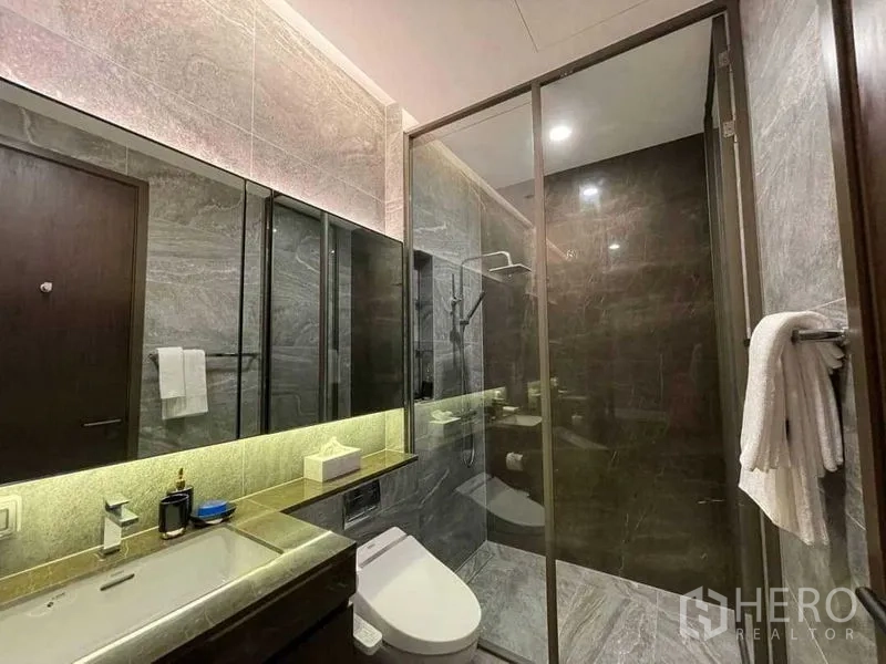 Condominium for rent in Khlong Toei, Bangkok - Modern bathroom featuring a glass‑enclosed rain shower, stone tiles and a backlit mirror vanity.
