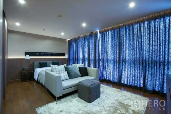 Condominium for sale in Sathon, Bangkok - Large bedroom with sitting area, wood floors and blackout curtains in a Bangkok condominium.