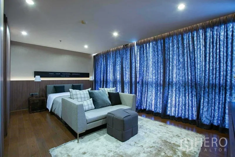 Condominium for sale in Sathon, Bangkok - Large bedroom with sitting area, wood floors and blackout curtains in a Bangkok condominium.