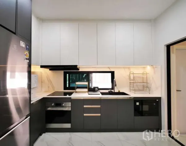 Detached House for rent or sale in Thanyaburi, Pathum Thani - Contemporary L-shaped kitchen with white upper cabinets, dark lower units, built-in oven and window over the sink.