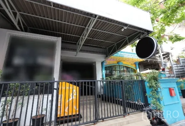 Shophouse for rent or sale in Watthana, Bangkok - Street-facing shophouse facade with metal canopy, round sign and gated entry surrounded by greenery.