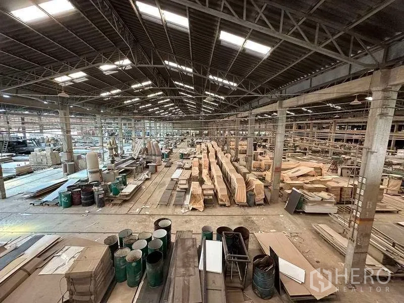 Factory for rent in Sai Noi, Nonthaburi - Large warehouse interior showing timber stacks and barrels beneath skylights in a metal-roofed hall.
