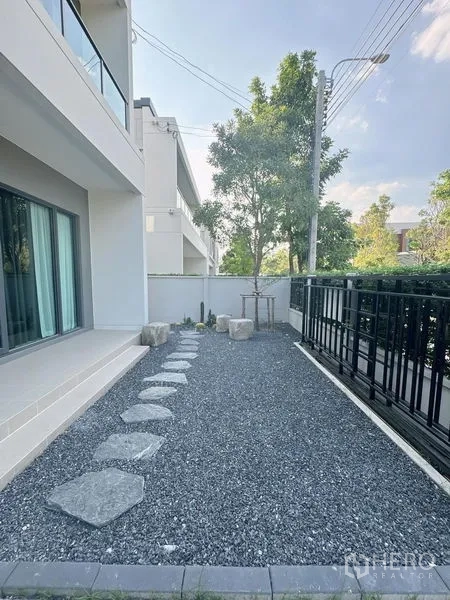 Detached House for rent or sale in Bang Phli, Samut Prakan - Side yard with gravel and stepping stones next to a modern two-storey house and black fence.
