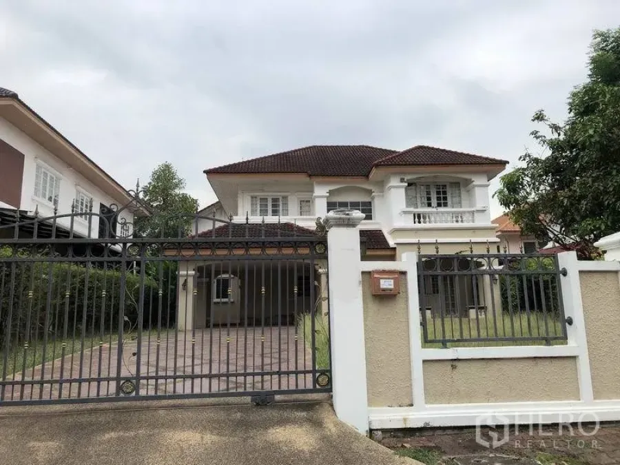 บ้าน ให้เช่าหรือขาย - บ้านเดี่ยวสองชั้นสีขาว หลังคากระเบื้องน้ำตาล รั้วเหล็ก ประตูทางรถเข้าและสนามหญ้าหน้าบ้าน ใต้ท้องฟ้ามีเมฆ