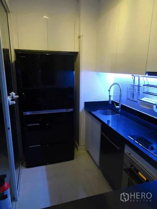 Condominium for rent or sale in Watthana, Bangkok - Galley kitchen view with glossy black fridge, under-cabinet lighting, sink and built-in oven.