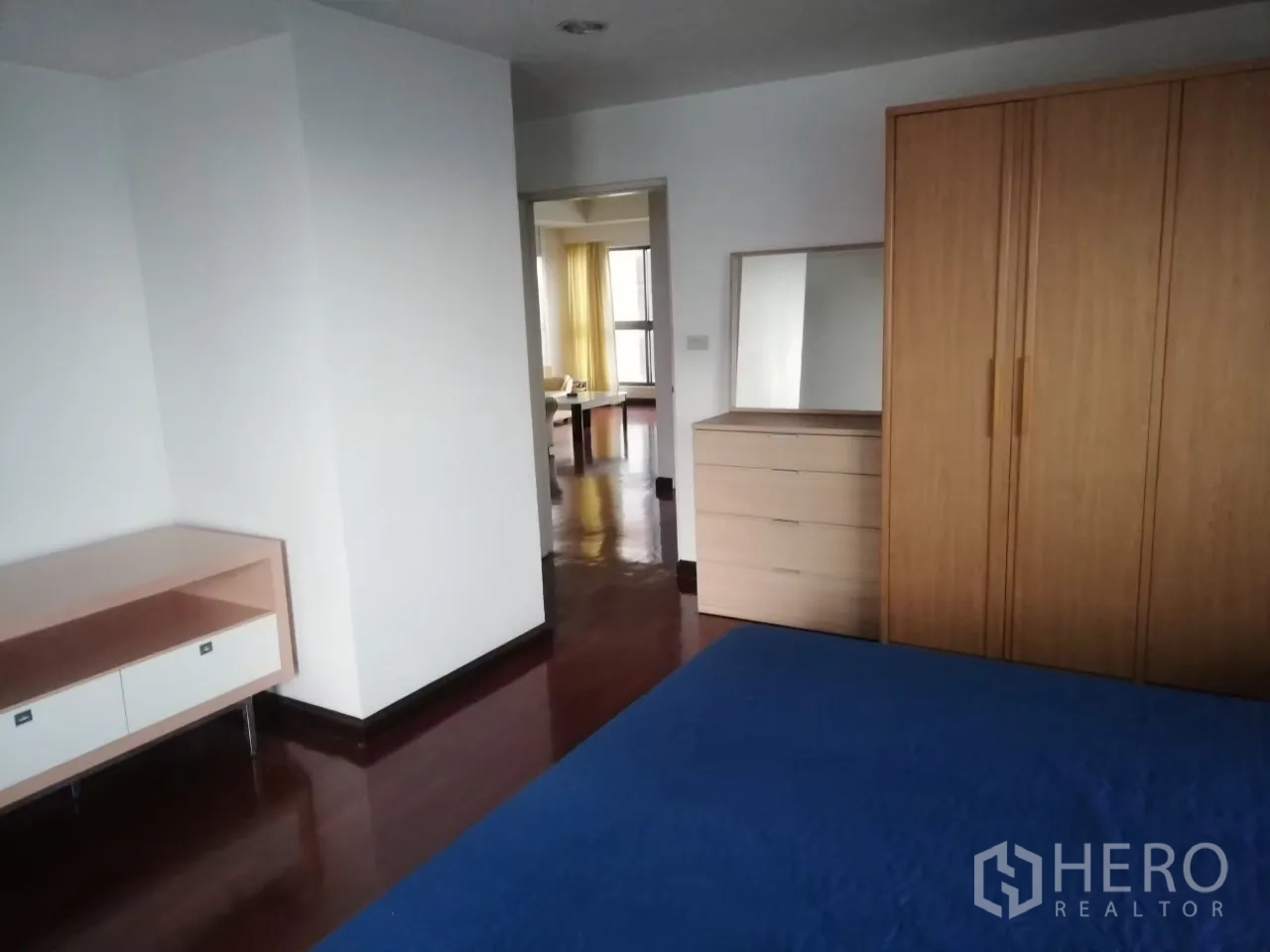 Condominium for rent in Phaya Thai, Bangkok - Primary bedroom with blue bedspread, wooden wardrobe and dresser, and polished hardwood floors leading to the living area.