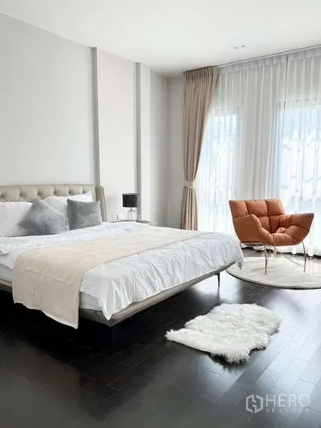 Detached House for rent or sale in Bang Phli, Samut Prakan - Bright master bedroom with tufted headboard, white bedding and an orange lounge chair beside floor-to-ceiling windows.