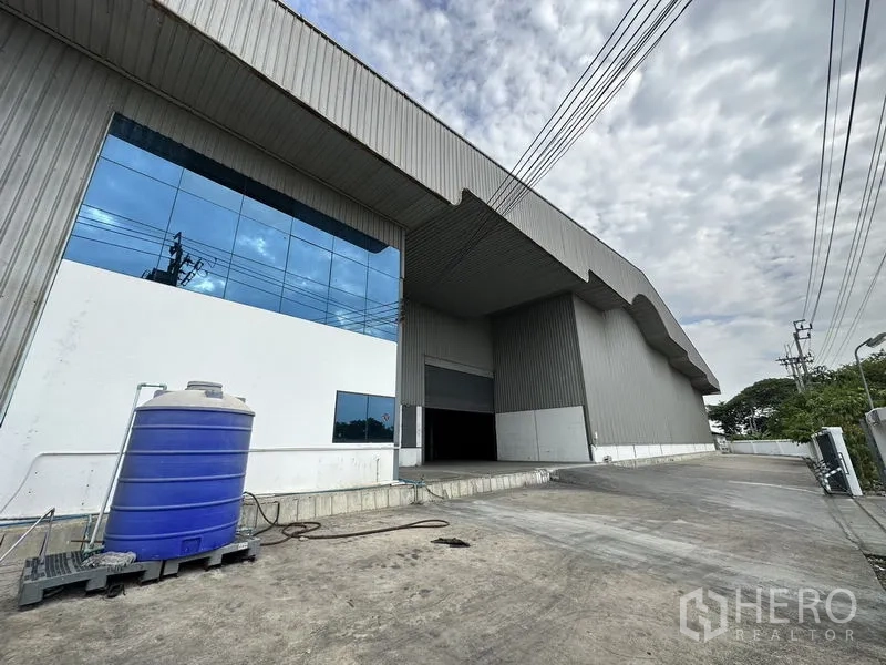 Warehouse for rent in Mueang Samut Prakan, Samut Prakan - Side perspective of the warehouse showing reflective glass office windows, loading dock and wide truck access lane.