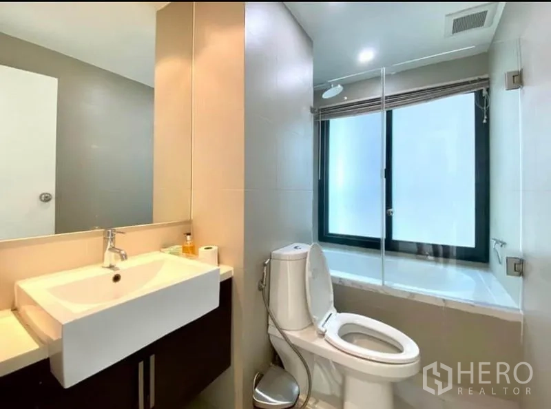 Condominium for rent in Khlong Toei, Bangkok - Modern bathroom with vanity, toilet, and glass-screened tub with shower.