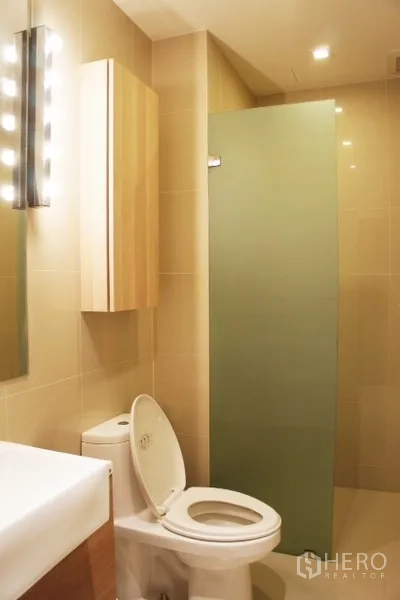 Condominium for rent in Phaya Thai, Bangkok - Modern bathroom with beige tiles, toilet, vanity and frosted glass shower screen.