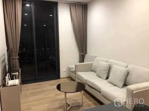 Condominium for rent in Khlong Toei, Bangkok - Cozy living room with light-gray sofa, round coffee table, wood floors, and sliding glass door with curtains showing city lights.