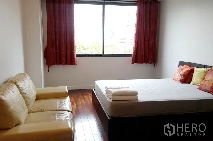 Condominium for rent in Khlong Toei, Bangkok - Spacious bedroom with a large window, red curtains, leather sofa, and green city view.