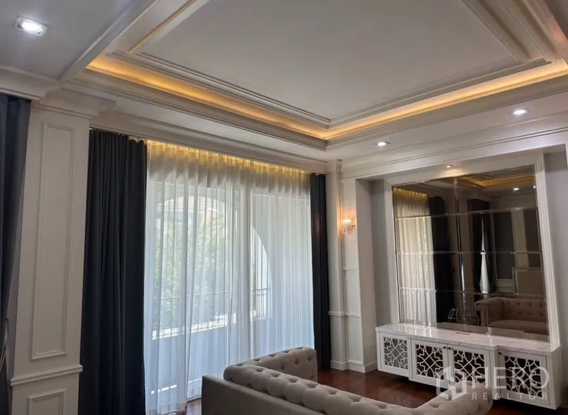 Detached House for rent or sale in Prawet, Bangkok - Elegant living room with coffered ceiling, warm cove lighting, sheer curtains, and a mirrored console.