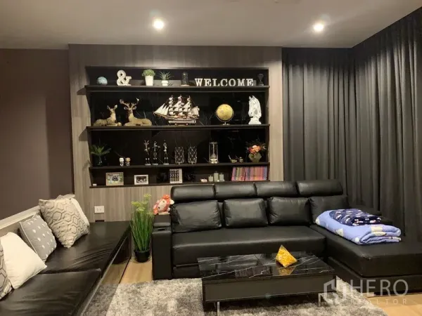 Townhouse for rent in Suan Luang, Bangkok - Contemporary living room with black leather sectional, built-in display shelves and dark drapes.