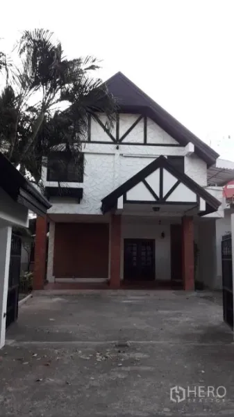 Detached House for rent in Yan Nawa, Bangkok - Front view of a two-storey detached house with gabled roof, Tudor-style trim, driveway and covered carport beside a palm tree.