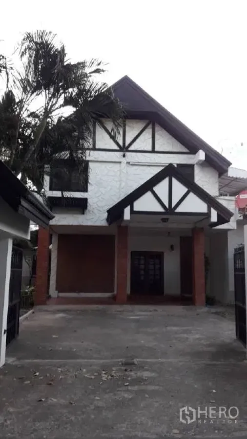 Detached House for rent in Yan Nawa, Bangkok - Front view of a two-storey detached house with gabled roof, Tudor-style trim, driveway and covered carport beside a palm tree.