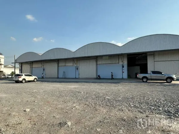 Warehouse for rent in Krathum Baen, Samut Sakhon - Row of warehouse bays with large shutter doors and a broad gravel forecourt for truck access.