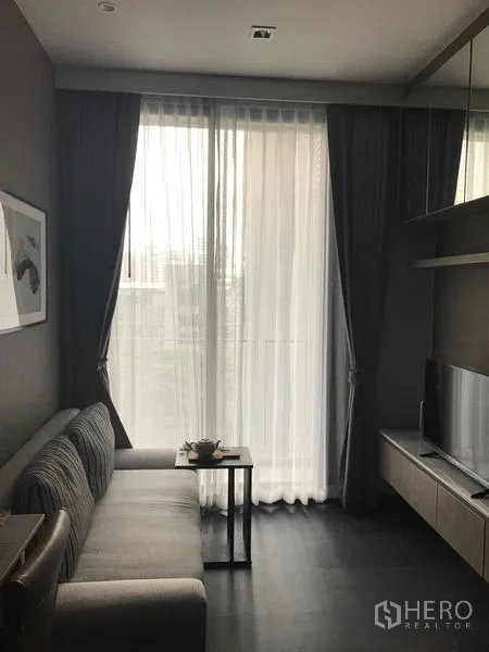 Condominium for rent in Watthana, Bangkok - Compact living area with gray sofa and sheer curtains opening to a tall sliding door.