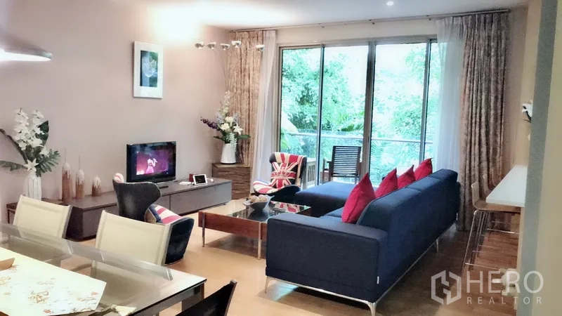 Condominium for rent in Bang Rak, Bangkok - Bright living room with navy sofa, red cushions, TV console and floor-to-ceiling windows to a leafy balcony.