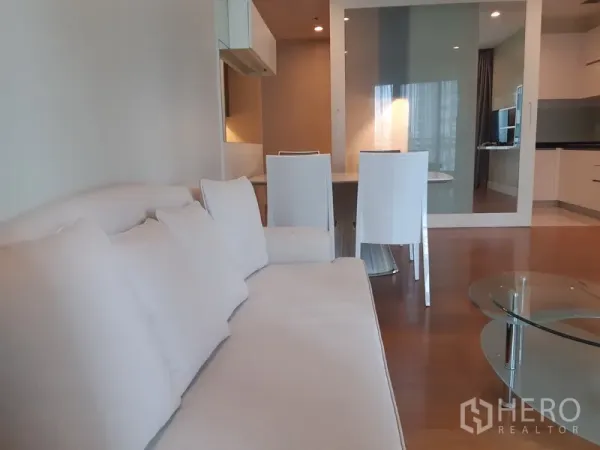 Condominium for rent in Khlong Toei, Bangkok - Open-plan lounge featuring a white sofa, glass coffee table, dining set, and mirrored wall beside large curtained windows.