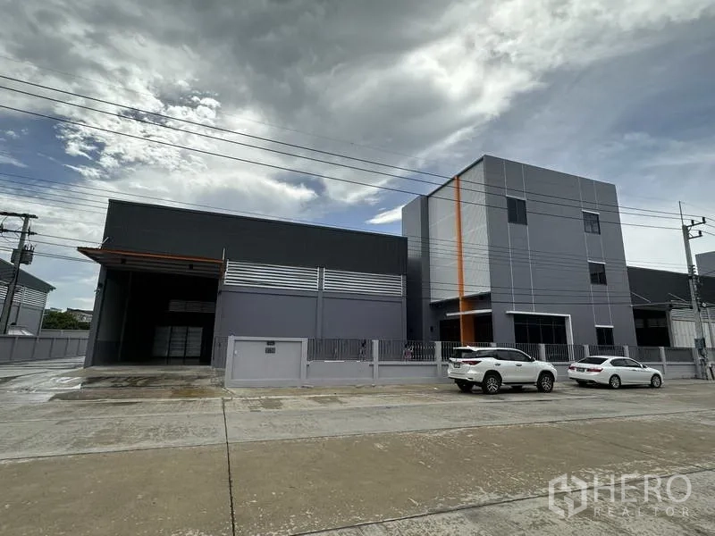 Factory for rent or sale in Lam Luk Ka, Pathum Thani - Front elevation of modern grey warehouse and office with parking spaces and perimeter fence.