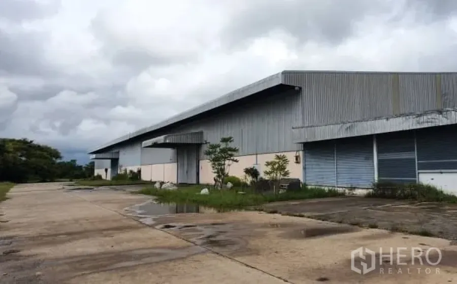 货栈 出租在Chaloem Phra Kiat, Nakhon Ratchasima - 大型金属外墙仓库外观，长屋顶、装卸月台与宽阔混凝土车道，天空多云。