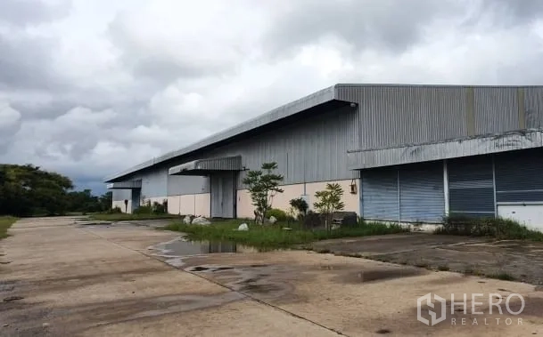 货栈 出租在Chaloem Phra Kiat, Nakhon Ratchasima - 大型金属外墙仓库外观，长屋顶、装卸月台与宽阔混凝土车道，天空多云。