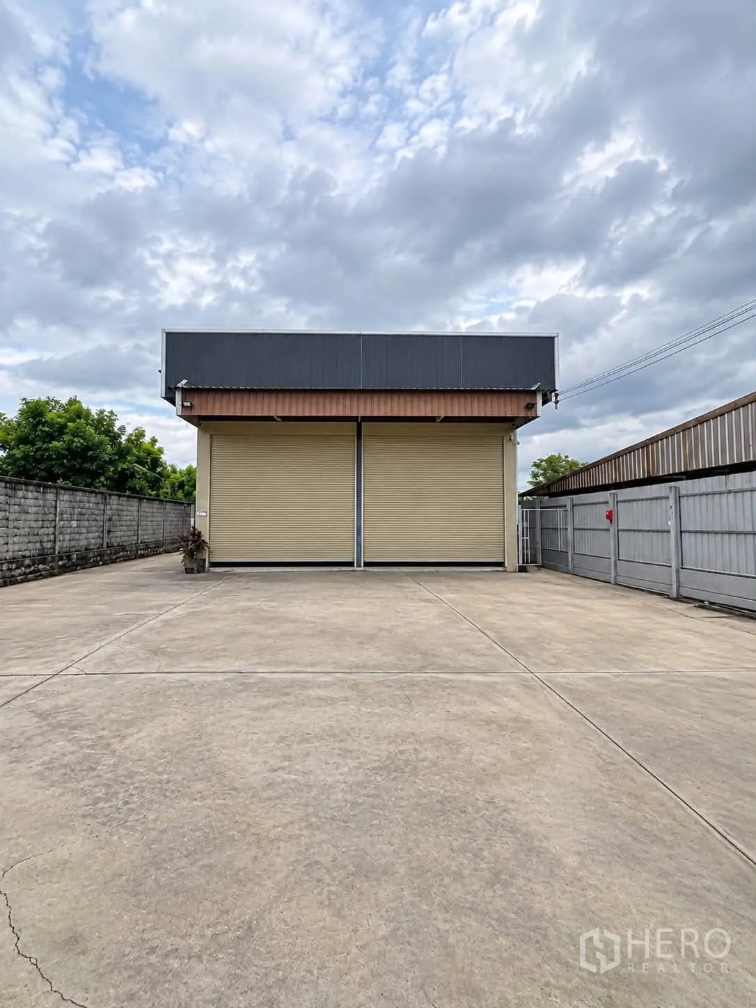 โกดัง for rent in Bang Kruai, Nonthaburi - ด้านหน้าโกดังพร้อมประตูม้วนคู่และลานคอนกรีตกว้าง มีรั้วและกำแพงล้อมรอบ