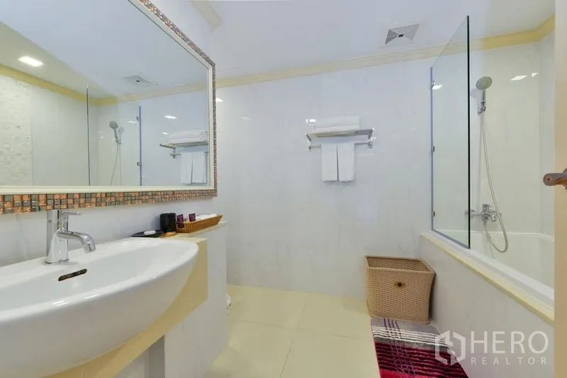 Apartment for rent in Khlong Toei, Bangkok - Bathroom vanity with wide mirror, storage basket, and glass-enclosed bathtub with shower.