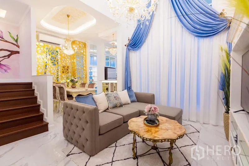 Townhouse for rent in Lat Phrao, Bangkok - Open-plan living and dining space with gray sofa, blue curtains, marble floors and a gold decorative partition.