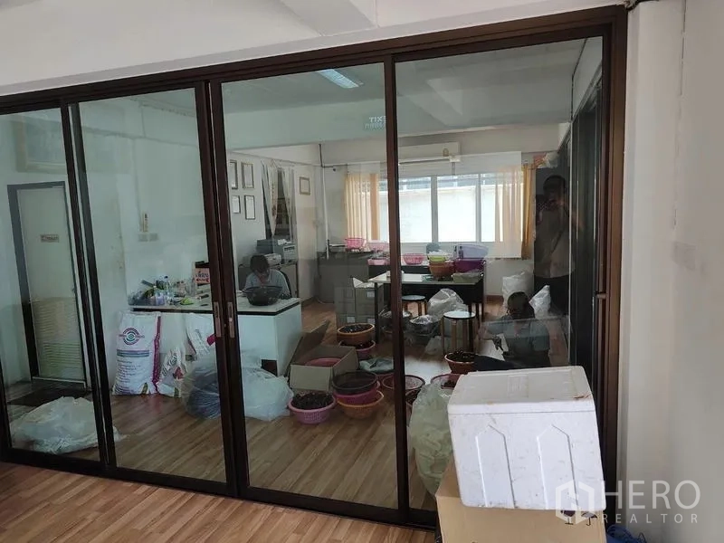 Factory for sale in Sai Noi, Nonthaburi - Glass-partitioned office area with desks and supplies on wood flooring inside the factory.