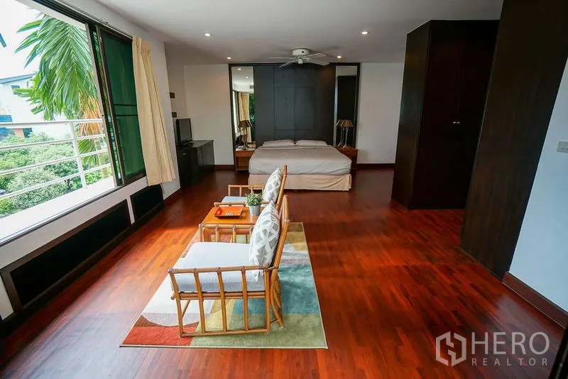 Apartment for rent in Watthana, Bangkok - Oversized master bedroom with seating area, wood flooring, and a wide window overlooking trees.