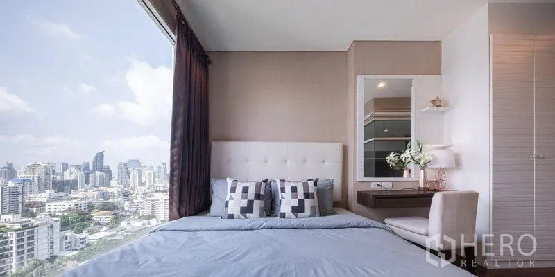 Condominium for rent or sale in Watthana, Bangkok - Bedroom with upholstered headboard, vanity desk and large window framing Bangkok skyline views.