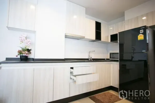 Condominium for rent in Watthana, Bangkok - Modern L-shaped kitchen with white cabinetry, black countertop and a full-size refrigerator.