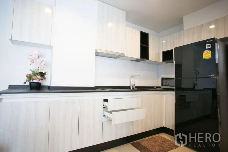 Condominium for rent in Watthana, Bangkok - Modern L-shaped kitchen with white cabinetry, black countertop and a full-size refrigerator.