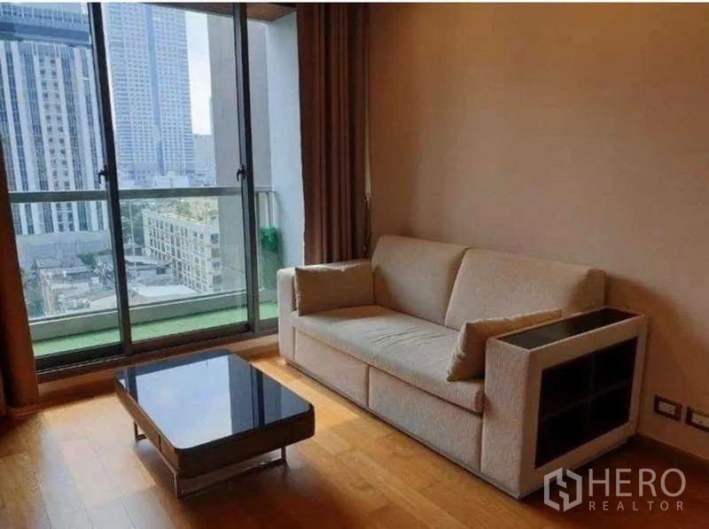公寓 出租在Bang Rak, Bangkok - 客厅采光充足，米色沙发与玻璃茶几，落地推拉门连通阳台，眺望沙吞天际线。