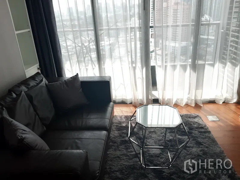 Condominium for rent in Khlong Toei, Bangkok - Bright living room with leather sofa, rug and floor-to-ceiling windows showing city views.