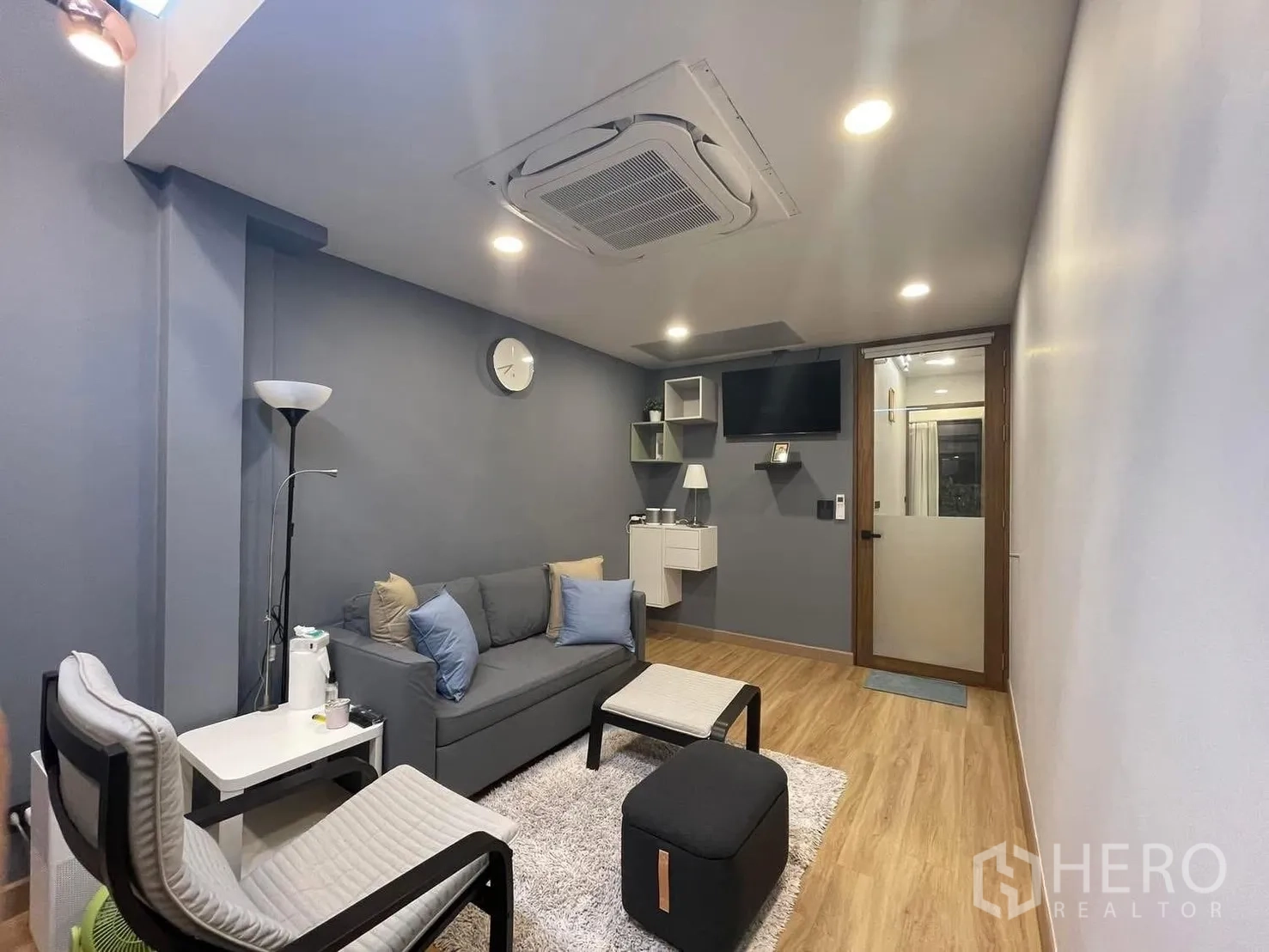 Home Office for rent or sale in Bang Kapi, Bangkok - Alternate view of the lounge with gray sofa, wall shelves, TV and warm lighting.