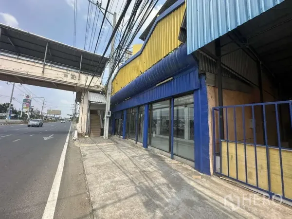 คลังสินค้า ให้เช่าในPak Kret, Nonthaburi - ด้านหน้าโกดังติดถนน ตัวอาคารสีเหลืองน้ำเงินพร้อมกระจกเต็มบานและสะพานลอยหน้าอาคาร