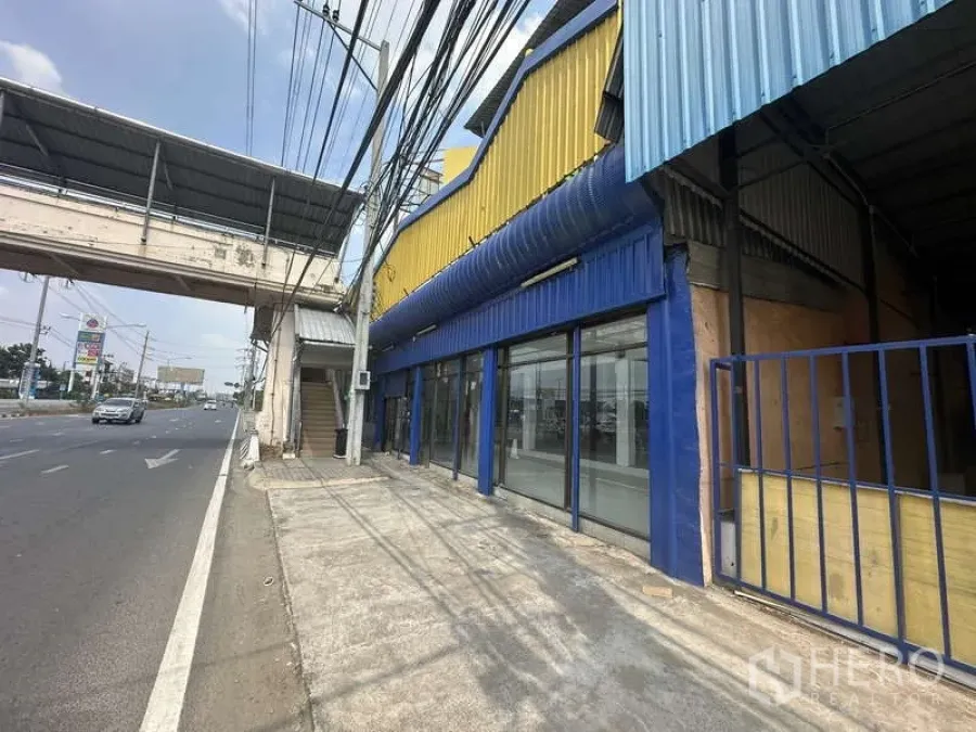 คลังสินค้า ให้เช่าในPak Kret, Nonthaburi - ด้านหน้าโกดังติดถนน ตัวอาคารสีเหลืองน้ำเงินพร้อมกระจกเต็มบานและสะพานลอยหน้าอาคาร