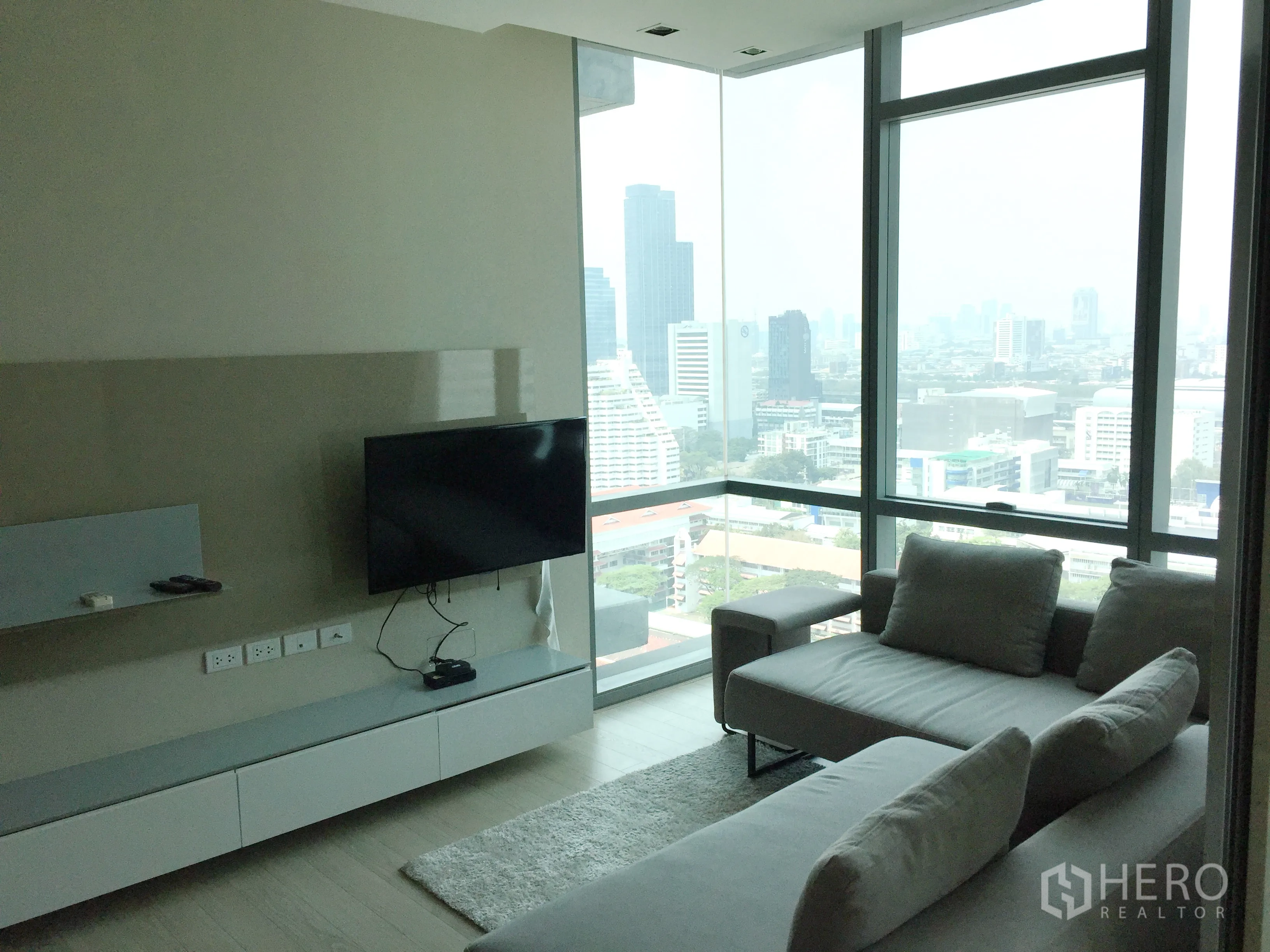 คอนโด ให้เช่าหรือขายในWatthana, Bangkok - ห้องนั่งเล่นกระจกเข้ามุม มุมมองวิวเมือง ทีวีติดผนัง และโซฟาสีเทา.