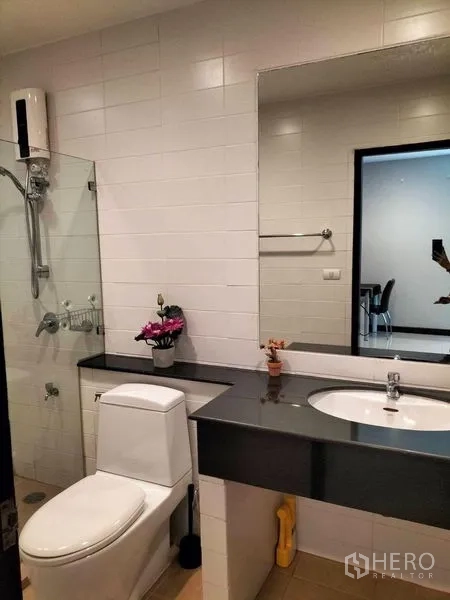 Condominium for rent in Watthana, Bangkok - Bathroom vanity with large mirror beside a glass shower and toilet.