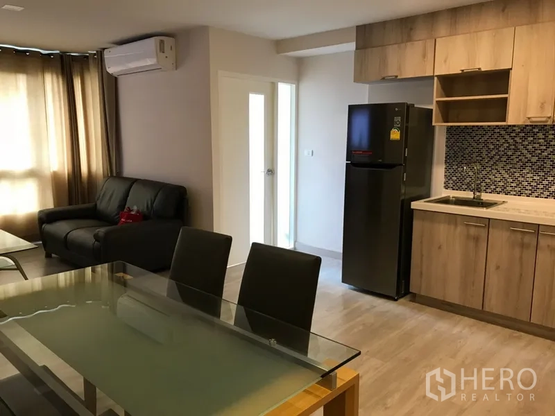 Condominium for rent in Watthana, Bangkok - Open‑plan living space with sofa, dining set, and kitchen featuring wood cabinets and a tall fridge.