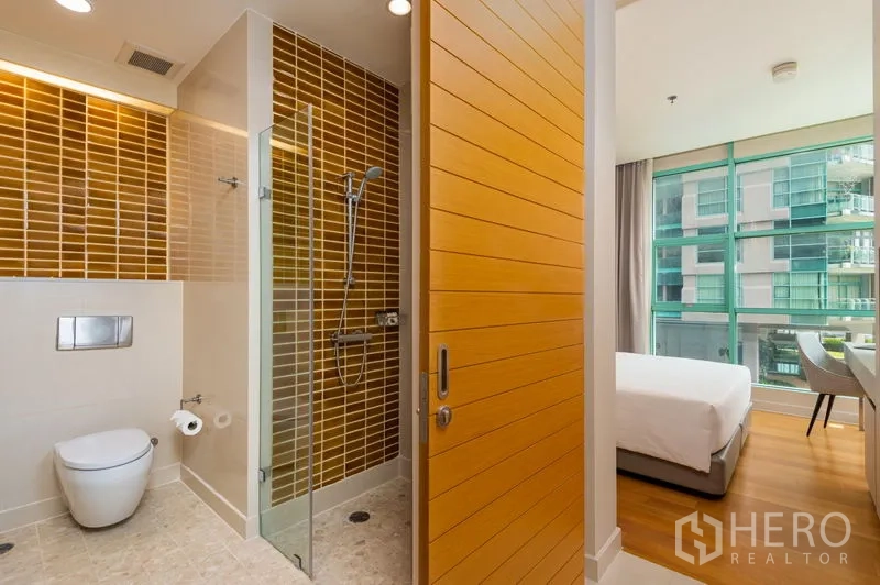 Condominium for rent in Bang Kho Laem, Bangkok - Modern bathroom with a walk-in glass shower, brown subway tiles, wall-hung toilet, and doorway to a bright bedroom with floor-to-ceiling windows.