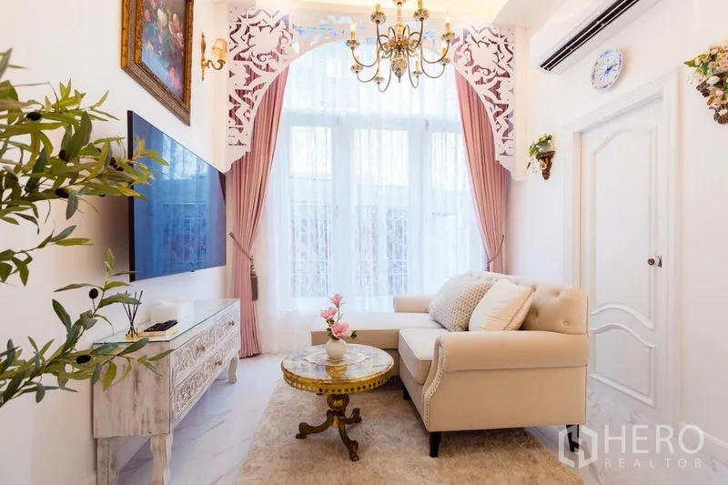 Townhouse for rent in Lat Phrao, Bangkok - Cozy lounge with cream L‑shaped sofa, wall-mounted TV, chandelier and pink drapes over tall windows.