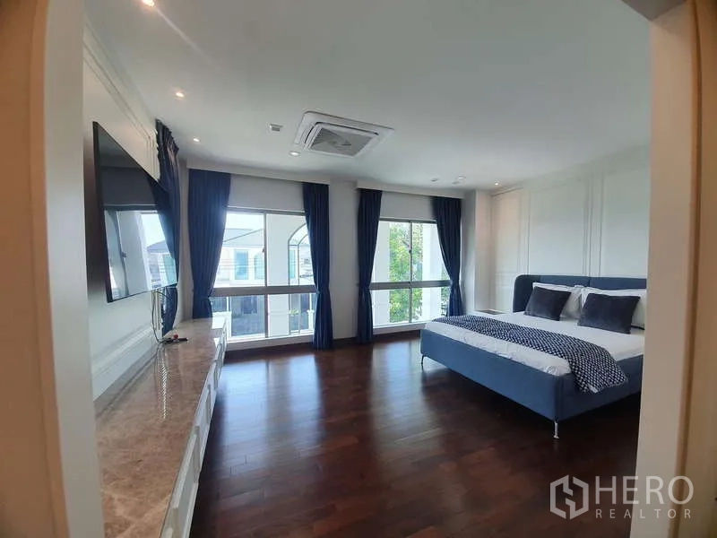 Detached House for rent or sale in Lat Krabang, Bangkok - Spacious master bedroom with hardwood floors, large windows, dark curtains and a TV wall.