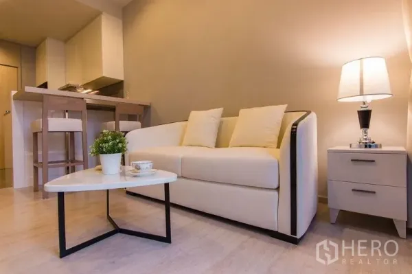 Condominium for sale in Watthana, Bangkok - Bright living area with cream sofa, side table lamp and dining bar in neutral tones.